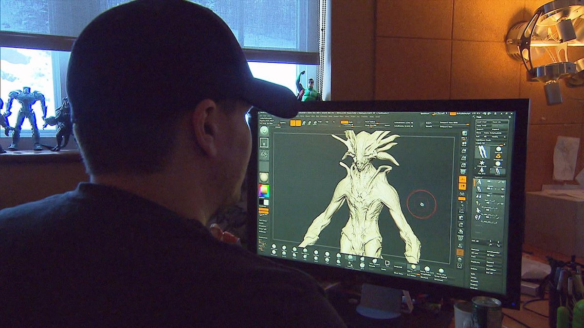 Un homme portant une casquette regarde un écran d'ordinateur où l'on peut voir une créature de jeu vidéo.