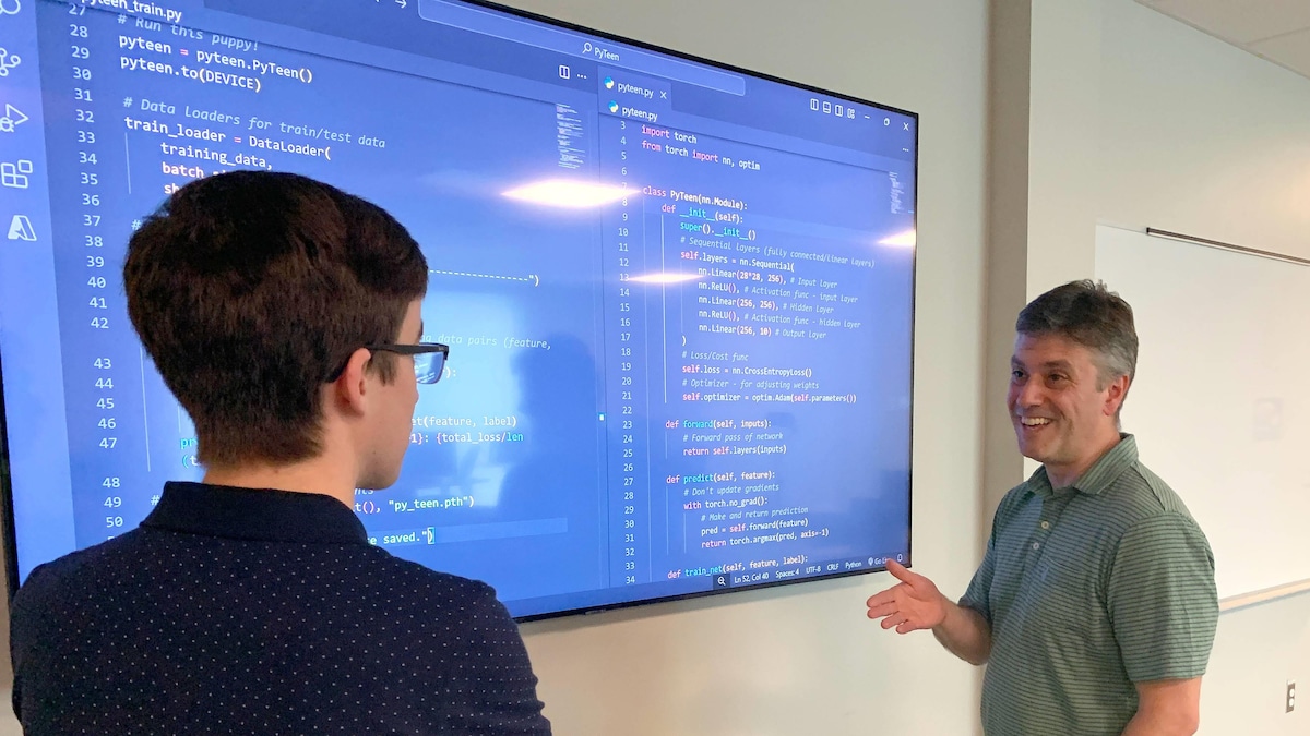 Un jeune homme debout, de dos, les bras croisés, regarde un professeur qui parle debout devant un écran géant qui présente des lignes de code sur un fond bleu.