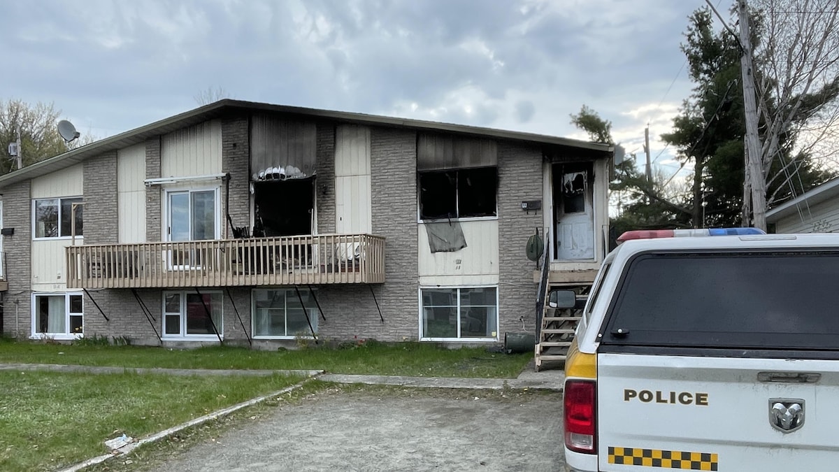Une voiture de la Sûreté du Québec est stationnée devant une maison endommagée par un incendie. Les fenêtres et la porte sont détruites et des traces de fumée apparaissent sur le devant de la maison.