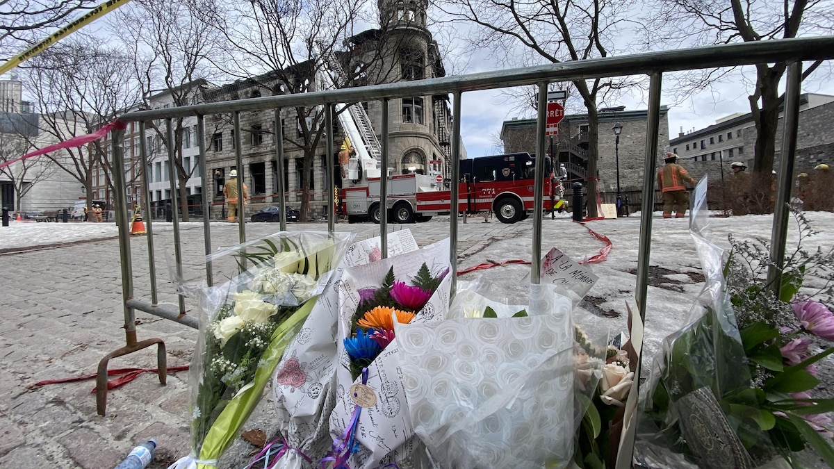 Des fleurs déposées près d'une barrière en métal devant un édifice incendié.