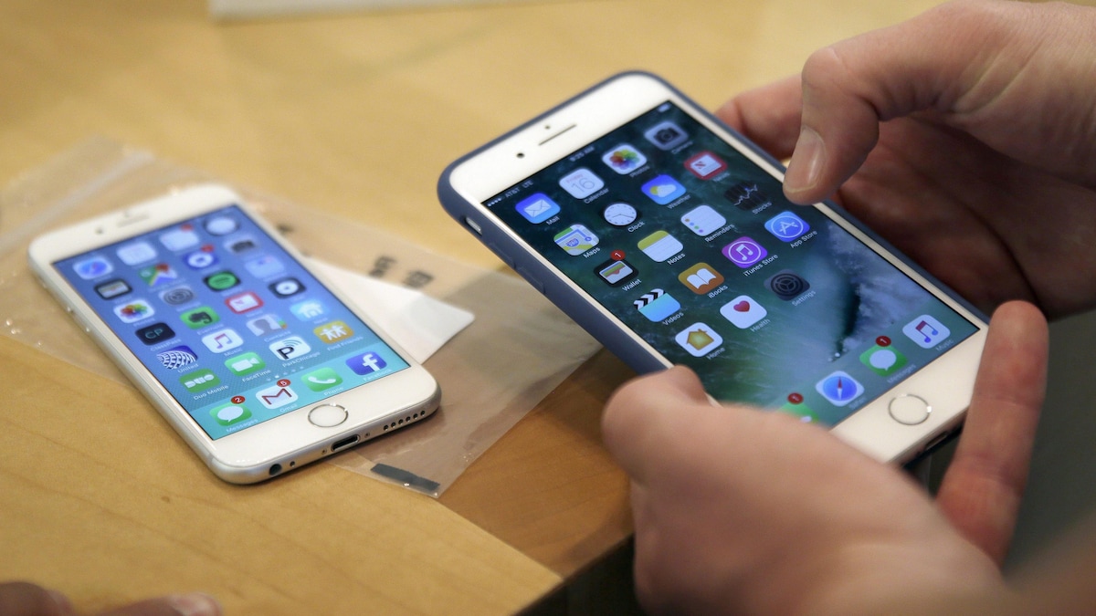 Plan rapproché des mains d'une personne manipulant un iPhone 7 Plus, alors qu'un iPhone 6 est posé sur une table en arrière-plan.