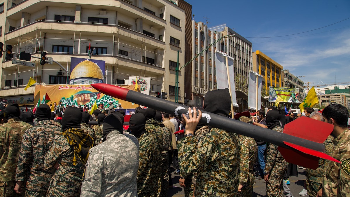 Des hommes en uniforme militaire marchent dans la rue. L'un d'eux tient une roquette sur son épaule.