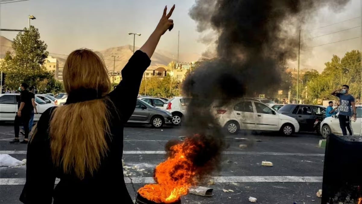 Une femme fait un signe de la main devant une voiture en flammes.