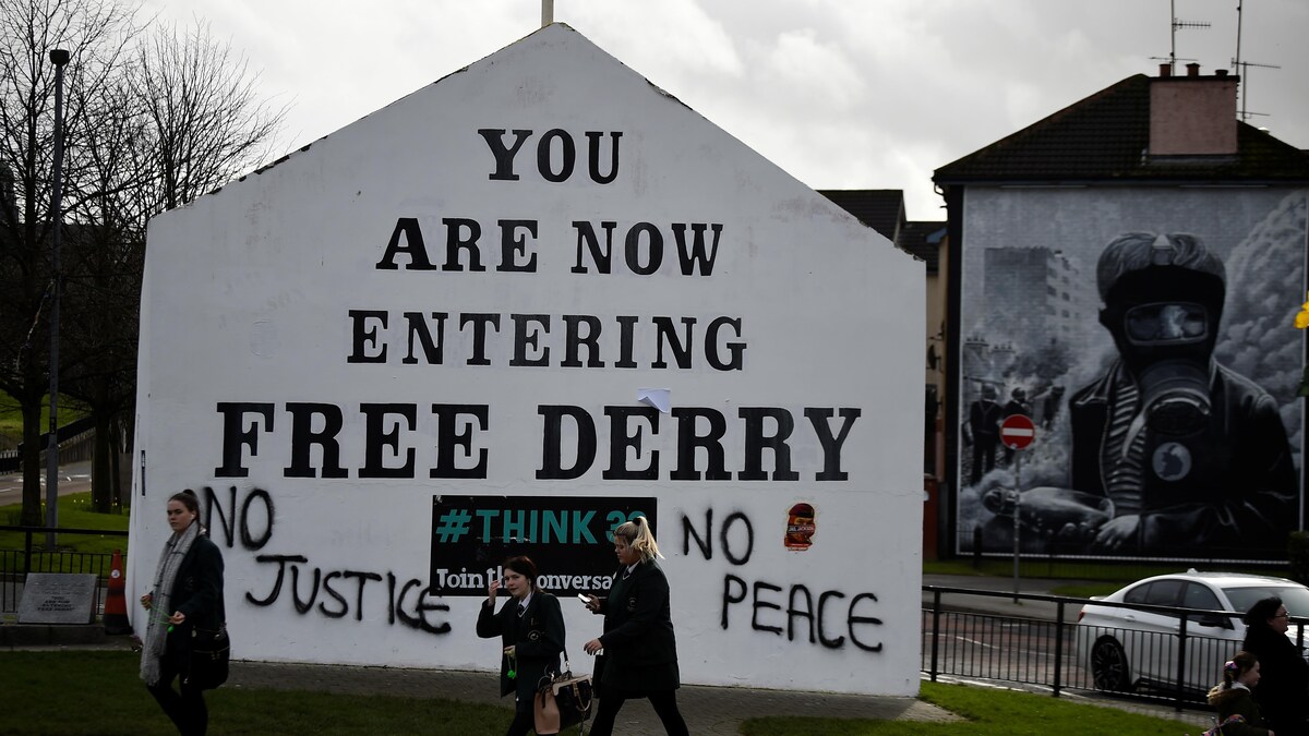 Des gens marchent devant un mur sur lequel il est écrit, en anglais, « Vous entrez maintenant dans Derry libre », avec des graffiti « No Justice » et « No Peace ».