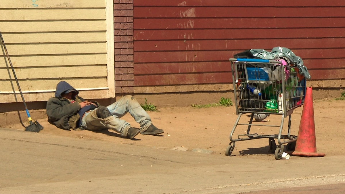 Un sans-abris dort couché contre un bâtiment près d'un panier d'épicerie rempli d'effets personnels. 