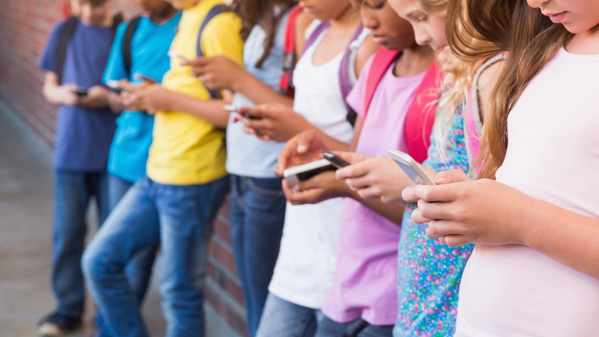 Des enfants portant des sacs à dos tiennent un appareil électronique dans leurs mains et le regardent. Ils sont accotés sur un mur de brique. 