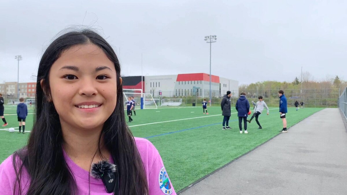Une adolescente sourit, debout sur les lignes de côté d'un terrain de football synthétique extérieur.