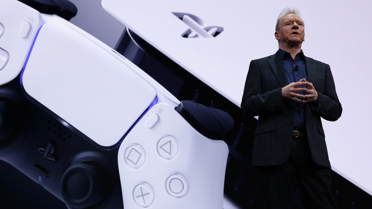 Un homme fait toucher les doigts de ses mains ensemble, et parle sur une scène devant une manette de jeu PlayStation et une console PlayStation 5. 