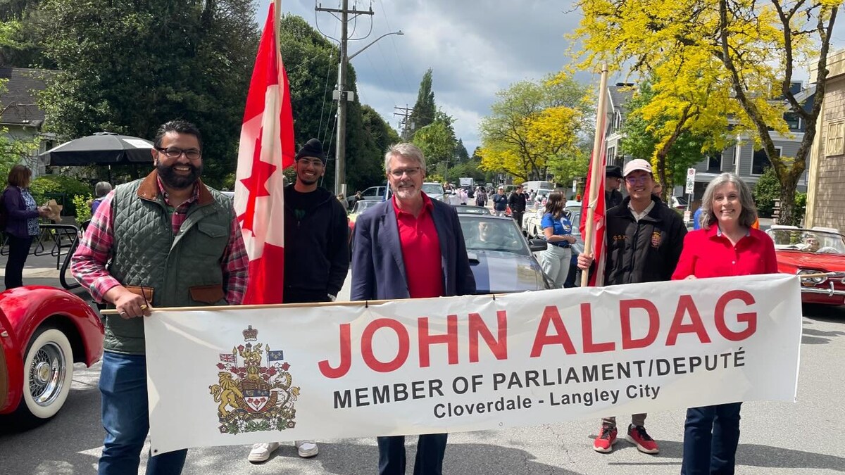 Un groupe de personnes derrière une banderole avec les armoiries du Canada où il est écrit John Aldag, member of parliament/député Cloverale-Langley.