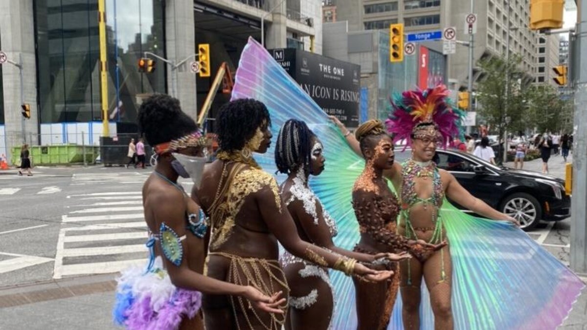 5 femmes portent des maillots deux pièces, des plumes et des paillettes de couleur, au coin d'une intersection achalandée du centre-ville de Toronto.