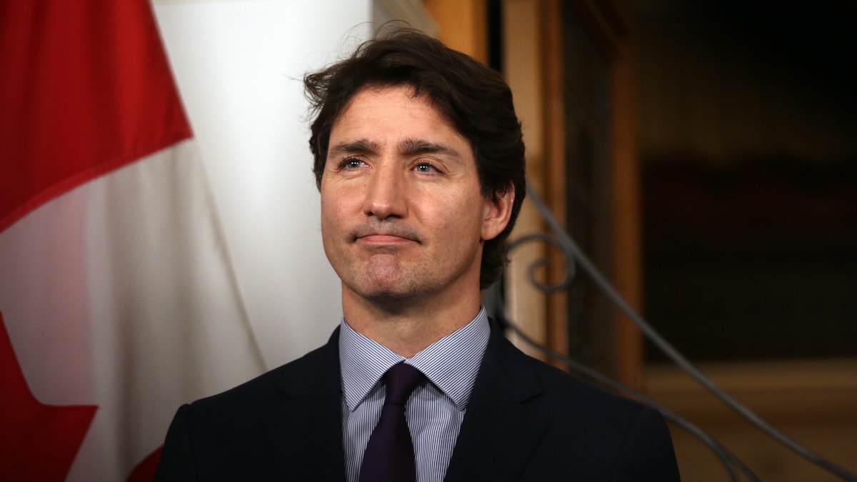 Devant un drapeau du Canada, Justin Trudeau regarde en semblant écouter un journaliste lui poser une question.