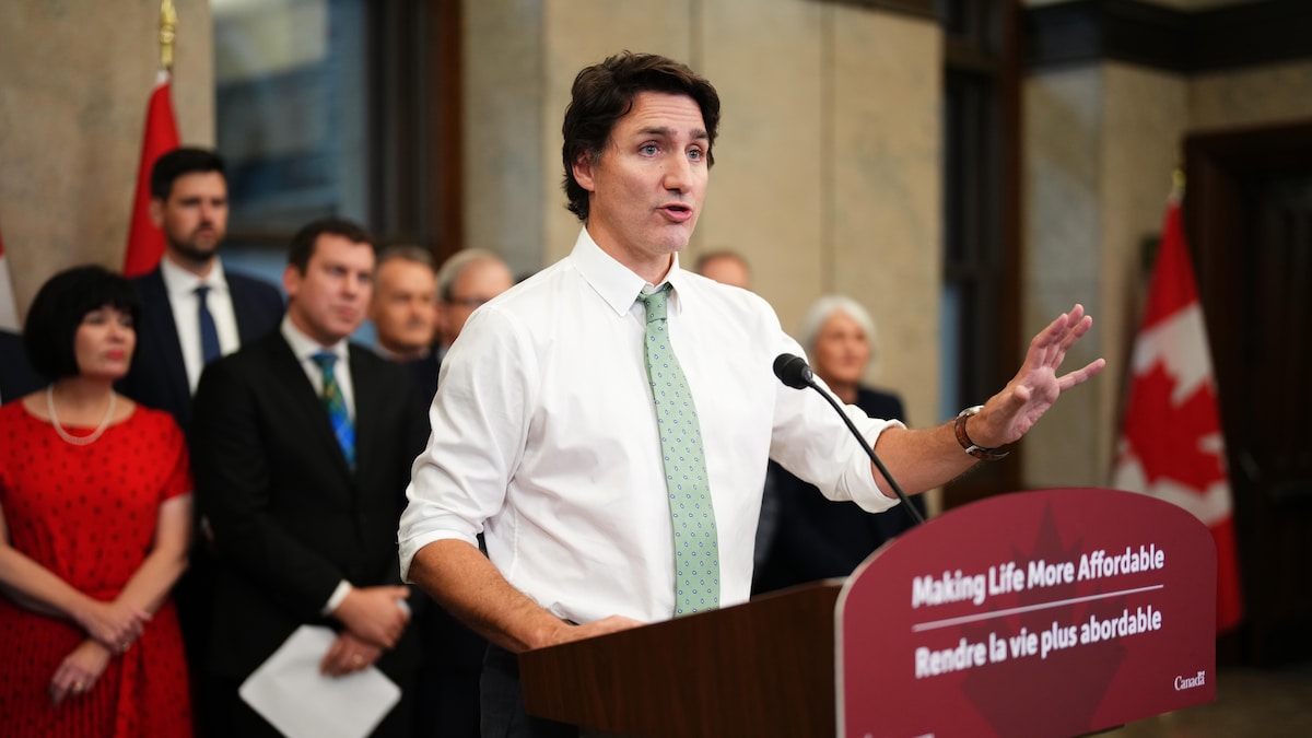 Justin Trudeau, en chemise blanche aux manches retroussées et cravate verte, parle au lutrin pendant que des députés sont debout derrière.