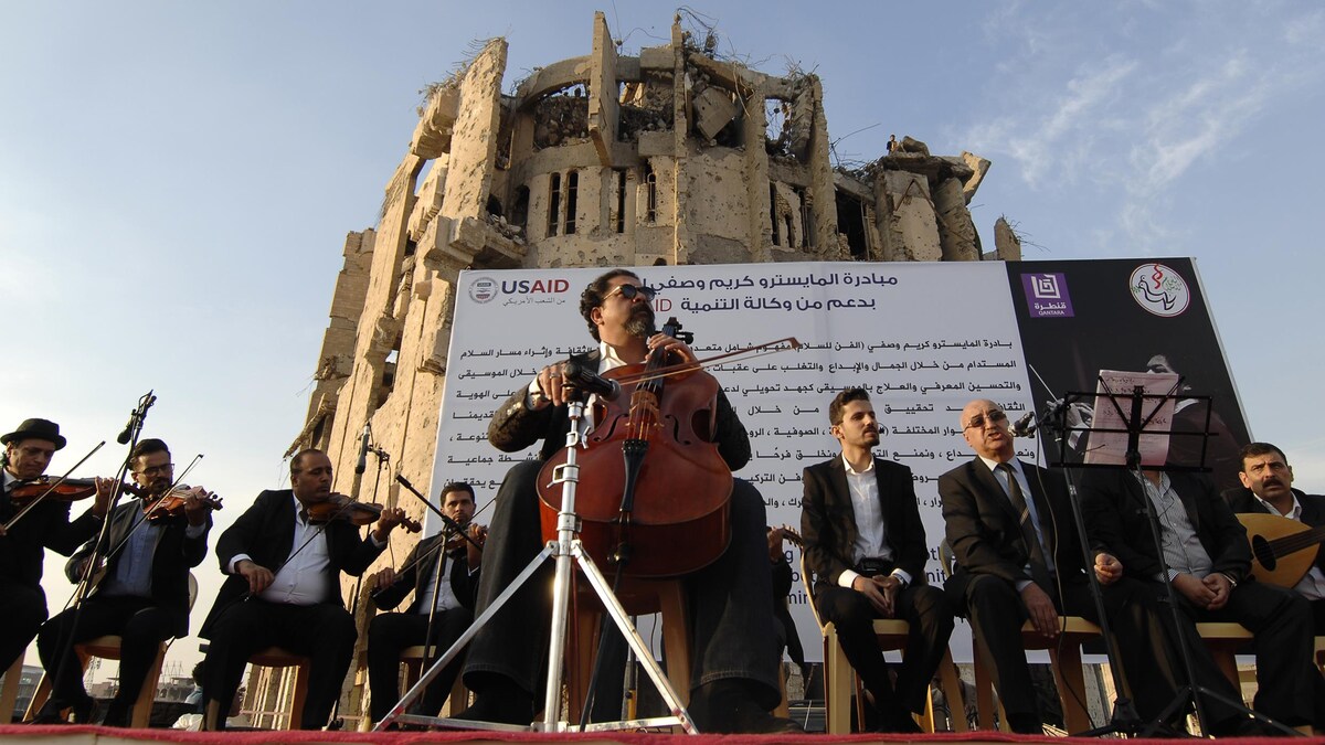 Le musicien est accompagné d'autres musiciens devant un bâtiment en ruines.