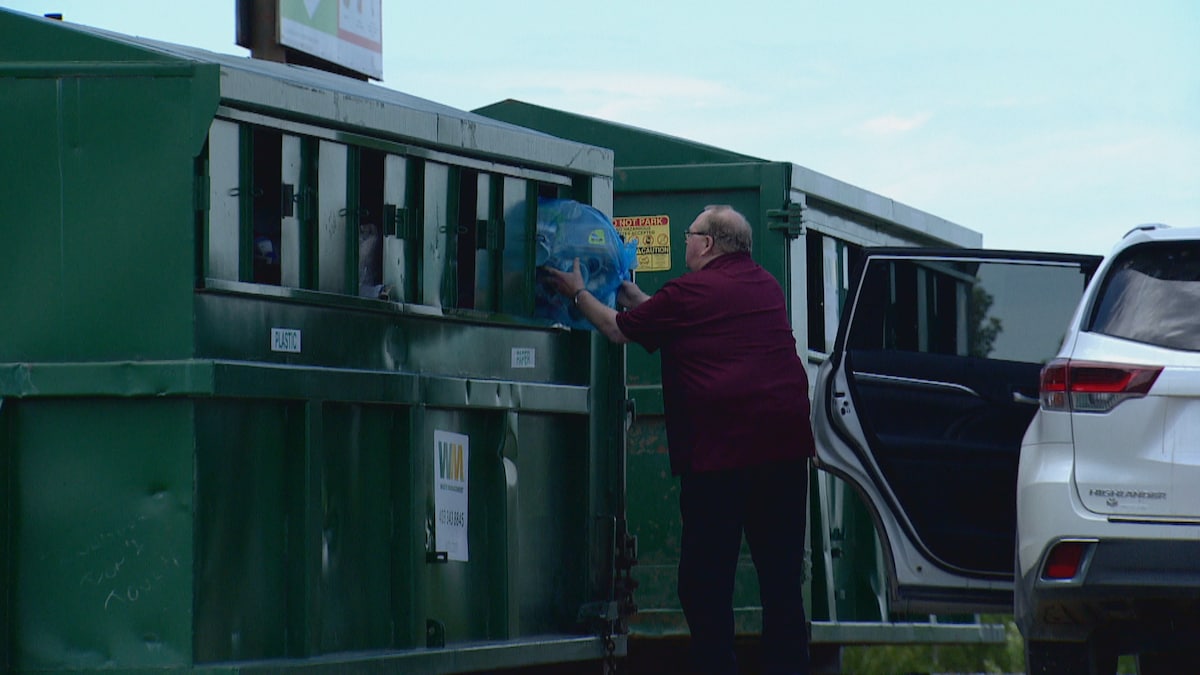 Un homme, à deux pas de sa voiture, dépose un sac de produits recyclabes dans un grand bac vert. 