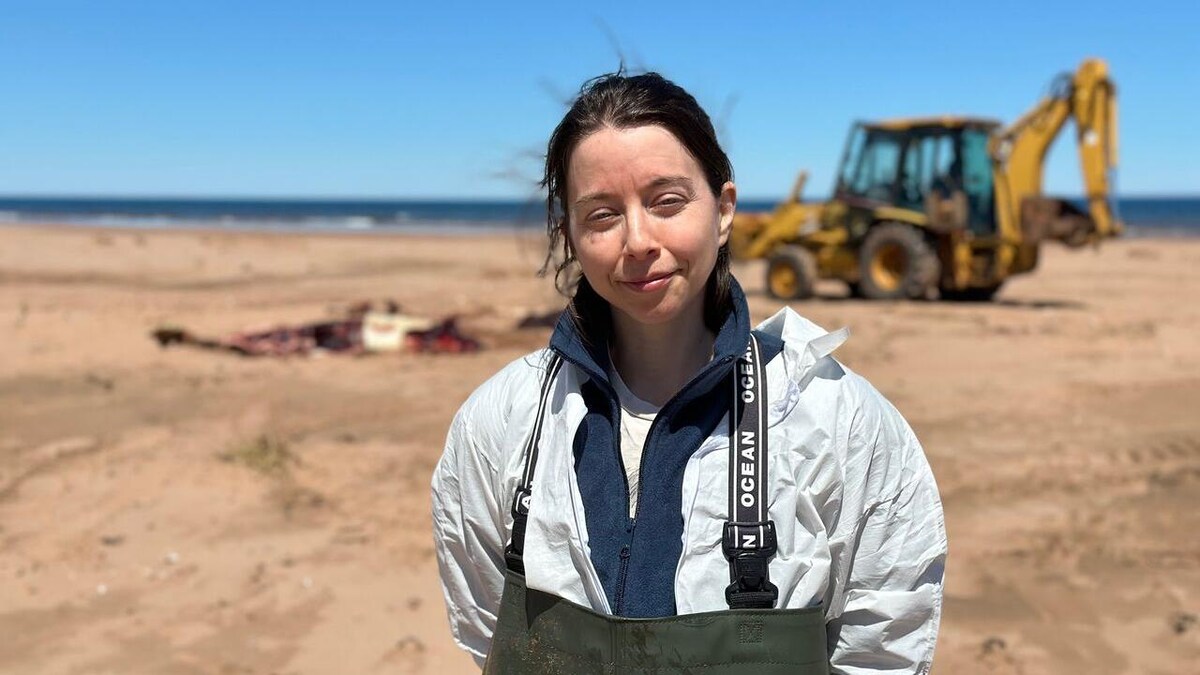 Une femme devant une carcasse de beluga sur une plage. Une pelle mécanique derrière elle. 