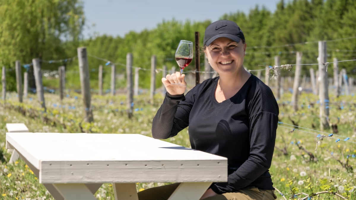 Une femme a un grand sourire alors qu'elle tient une coupe de vin et est assise à une table en plein milieu d'un vignoble.