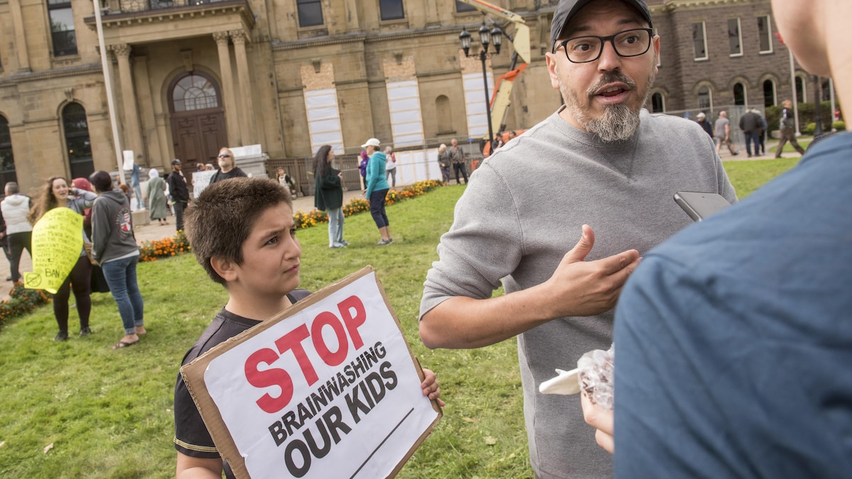 Deux personnes discutent près d'un enfant tenant une pancarte sur laquelle il est écrit : « Arrêtez de laver le cerveau de nos enfants ».