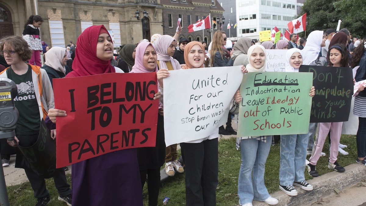 Des jeunes filles et une femme adulte portant des hijabs sont en rang sur la chaîne de trottoir et montrent des pancartes sur lesquelles ont lit des messages tels « J'appartiens à mes parents » et « Laissez l'idéologie hors des écoles ».