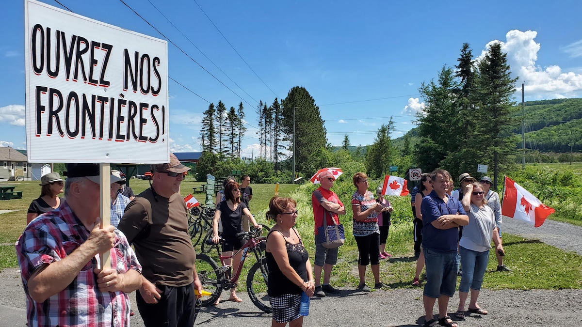 Un homme tient une pancarte « Ouvrez nos frontières! », il y a une dizaine de personnes à côté de lui et certains tiennent des drapeaux du Canada. 
