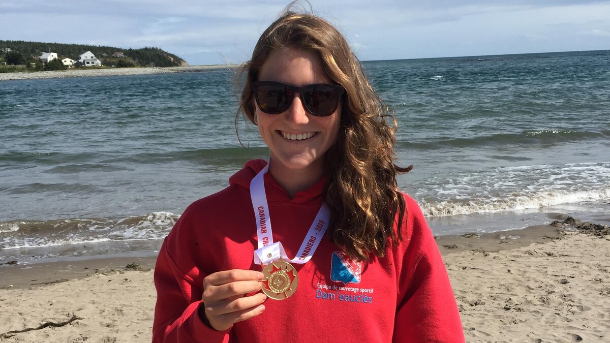 Manuelle Charbonneau est debout sur la plage, devant la mer, et sourit en tenant la médaille d'or qu'elle porte au cou. 