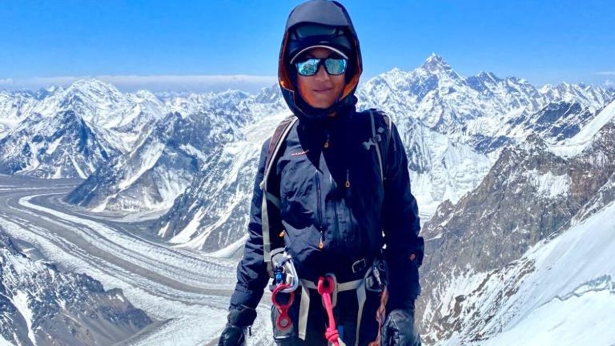Une alpiniste attachée par des harnais regarde la caméra. On voit derrière elle des sommets enneigés