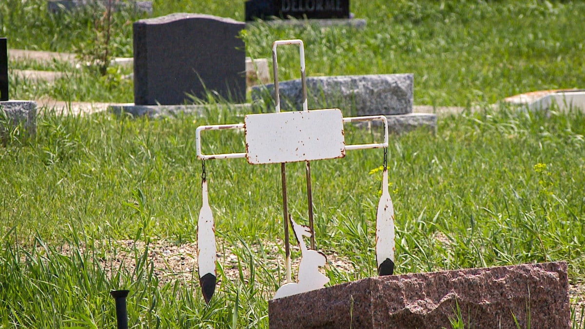 Une croix blanche de métal avec des plumes est postée devant une tombe.