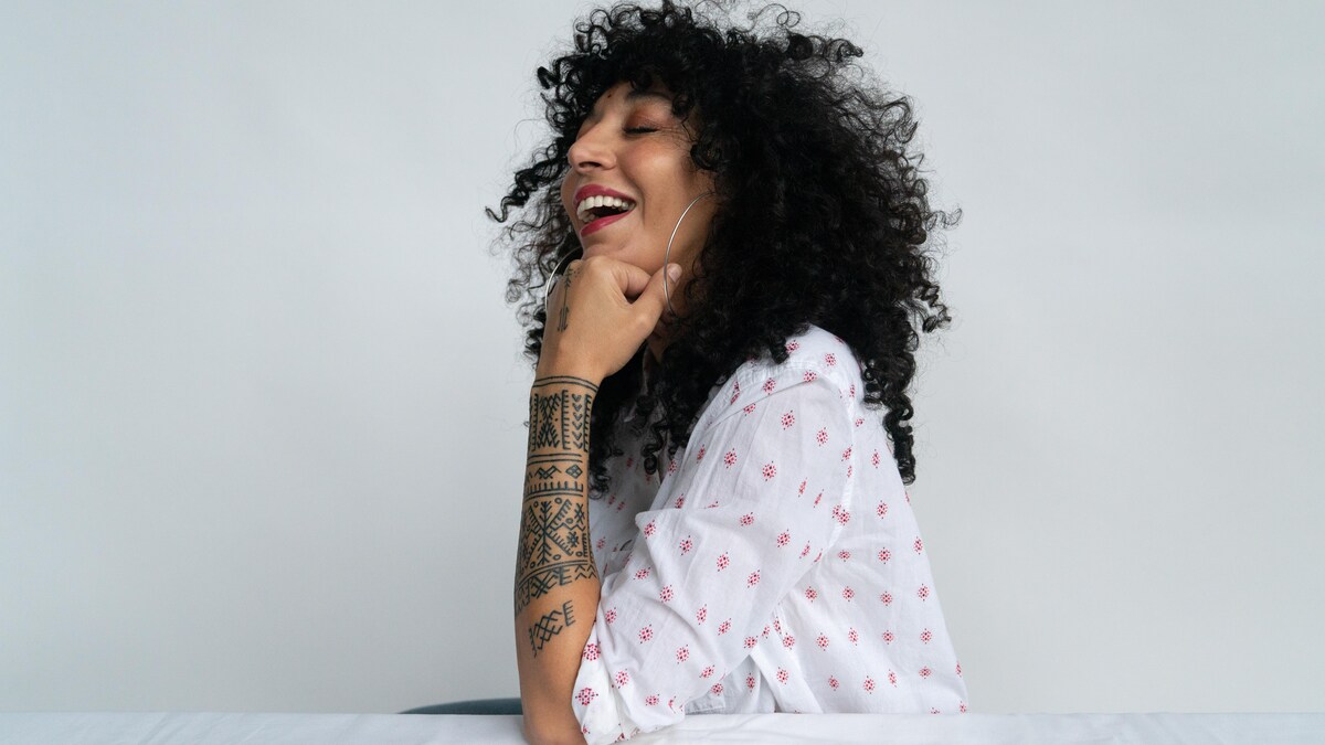 Maya est assise, de profil, derrière une table blanche. Elle rit, sa tête encadrée de longs cheveux bouclés noirs appuyée sur son poing. Son bras est recouvert de tatouages amazigh. 