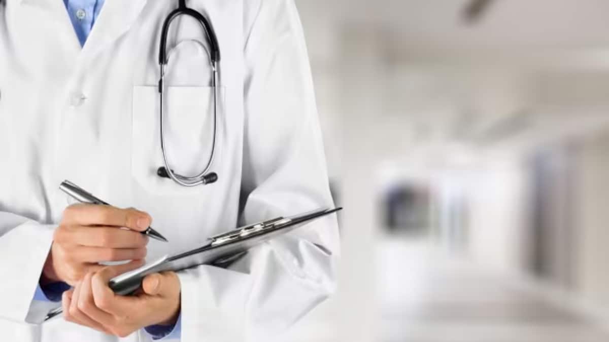 Un médecin dans un couloir d'hôpital s'apprête à écrire sur un document. Il porte un stéthoscope autour du cou.