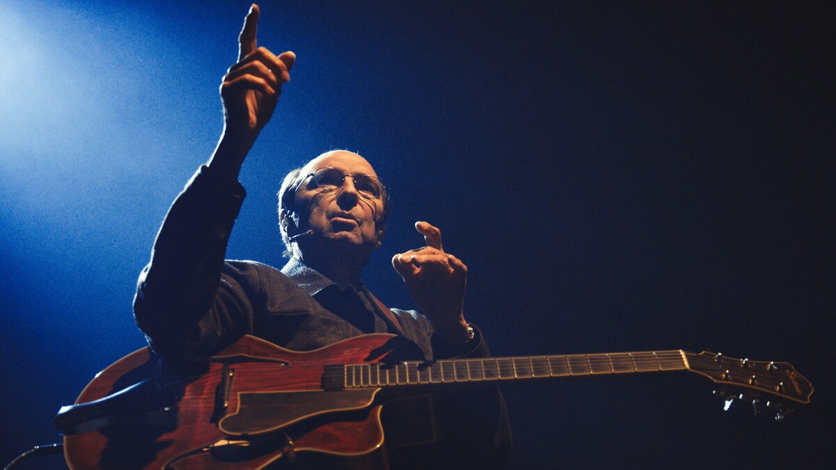 Michel Rivard avec sa guitare qui pointe devant lui.                                