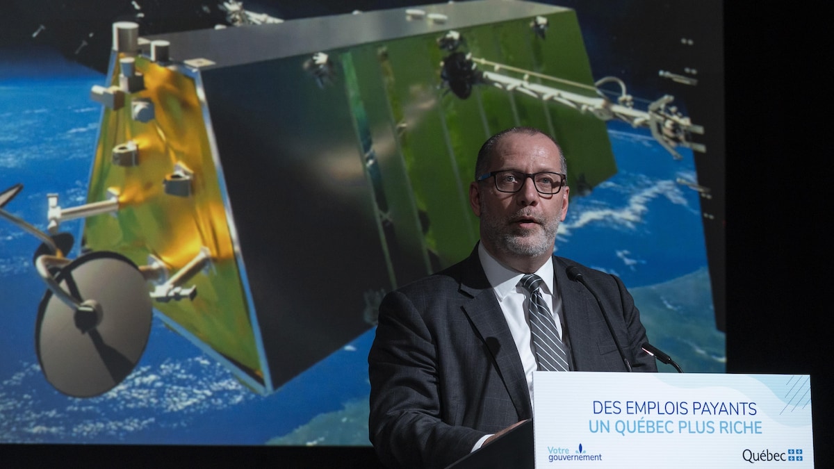 Mike Greenley, derrière un lutrin durant un point de presse, et devant une image d'un satellite dans l'espace.