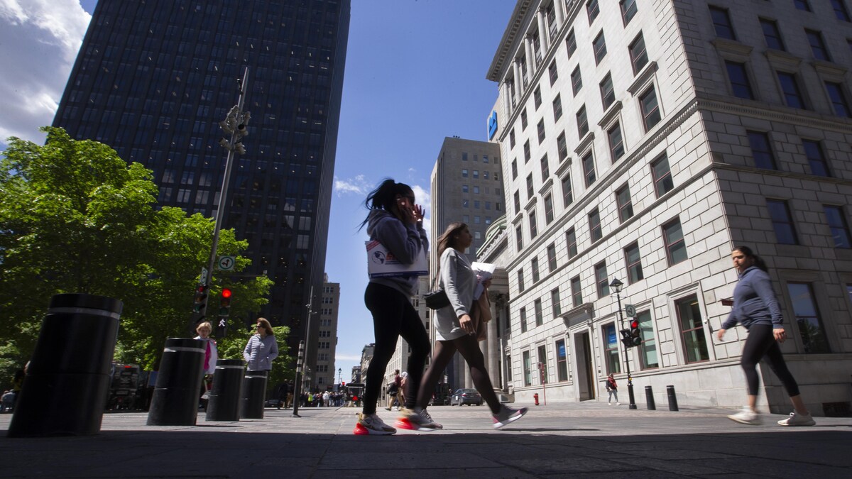 On voit des passants, au premier plan une jeune femme, qui marchent sur la place d'Armes, au centre-ville de Montréal. En arrière-plan, on voit les édifices emblématiques, dont celui de la BMO.