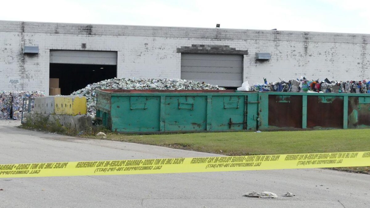 Un ruban définissant le périmètre d'une enquête policière s'étend à l'entrée d'un entrepôt en brique devant lequel se trouvent des conteneurs remplis de matières recyclables.