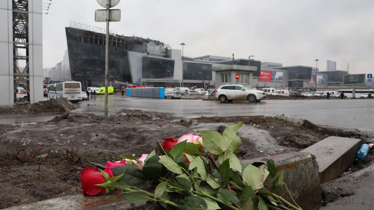 Un bouquet de rose est déposé sur une rambarde de béton devant un terrain boueux, une route, un bâtiment incendié et un ciel gris.