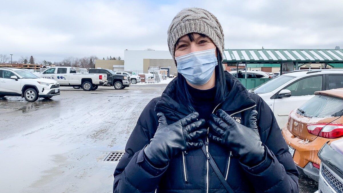 Une femme portant un masque s'adresse à la caméra dans le stationnement d'une épicerie.