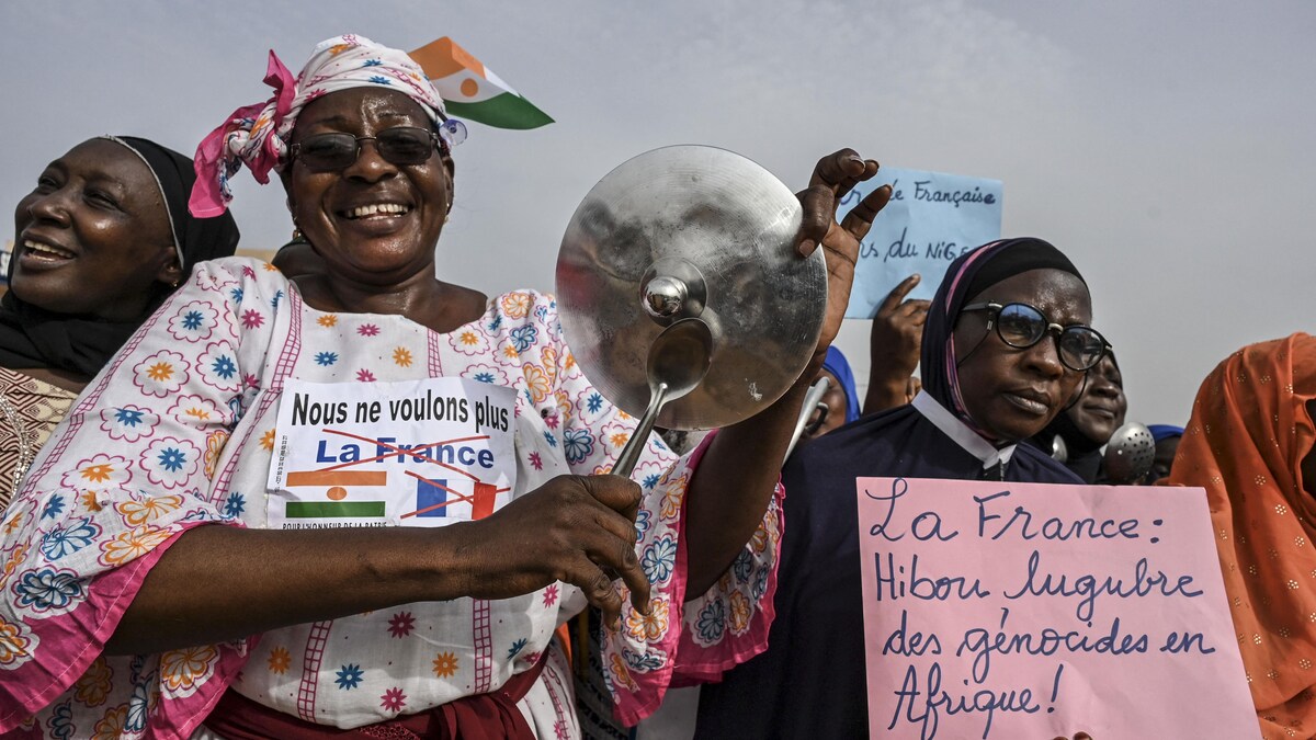Une femme souriante frappe sur le couvercle d'une casserole. Elle a une affiche qui dit : Nous ne voulons plus la France.
