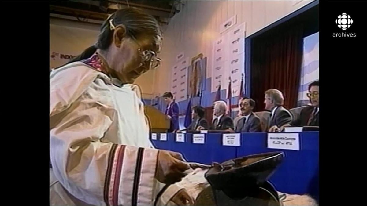 Cérémonie du feu d'une femme inuit. En arrière-plan, le premier ministre Brian Mulroney et les représentants autochtones sont assis au long d'une table pour signer l'accord.
