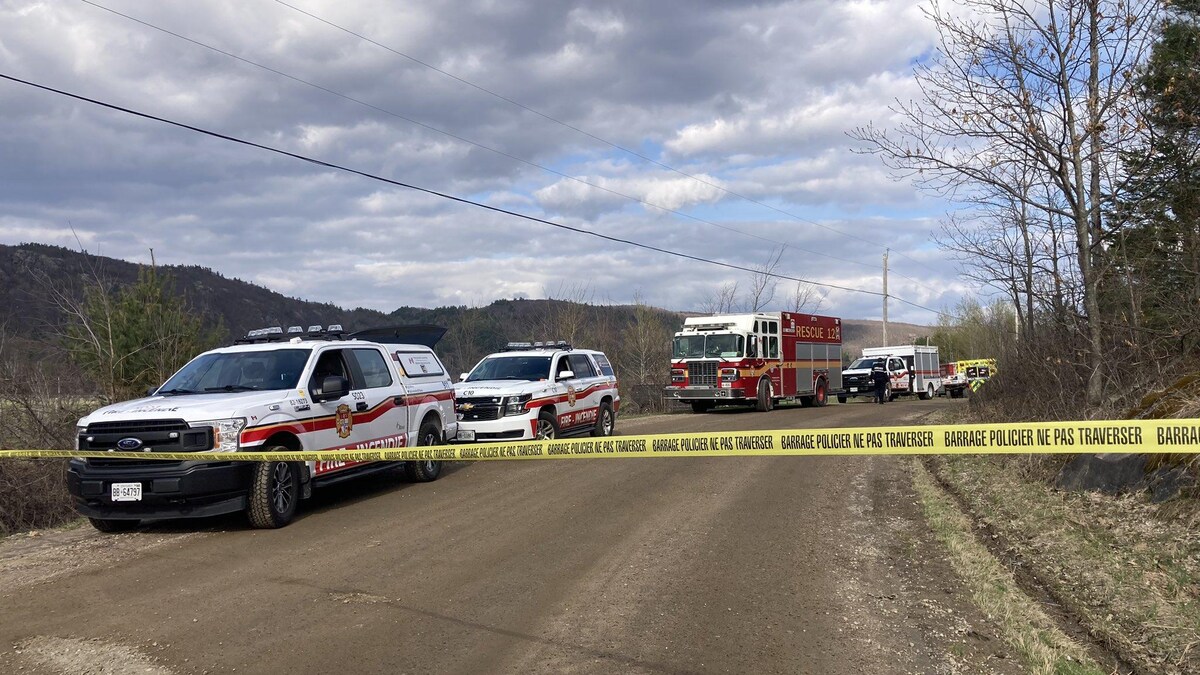 Deux camionnettes et un camion d'un service de sécurité incendie sont stationnés un à la suite de l'autre sur le bord d'une route, derrière un ruban jaune sur lequel il est écrit « Barrage policier, ne pas traverser ».