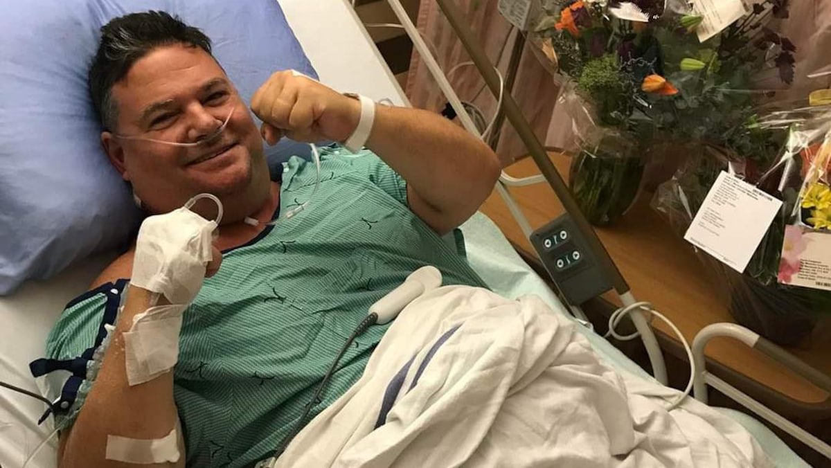 Un homme couché dans un lit d'hôpital après une opération sourit et fait un signe combatif avec ses deux poings serrés et levés comme s'il était un boxeur.