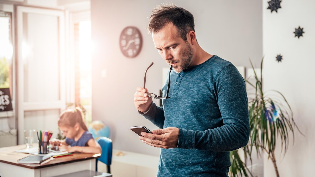 Un père regarde son téléphone avec un air inquiet pendant que sa fille fait ses devoirs en arrière-plan.