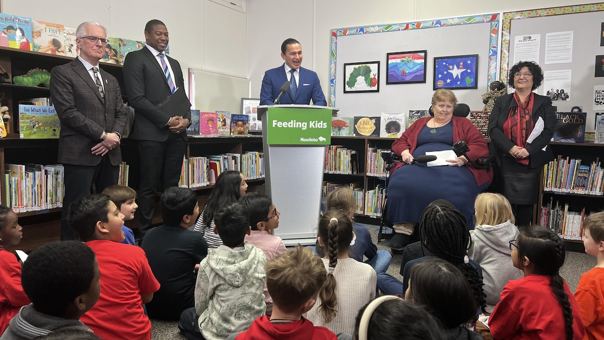 Le premier ministre du Manitoba, Wab Kinew, accompagnée des ministres Nello Altomare et Jamie Moses tiennent une conférence de presse dans la bibliothèque d'une école devant des enfants assis par terre.
