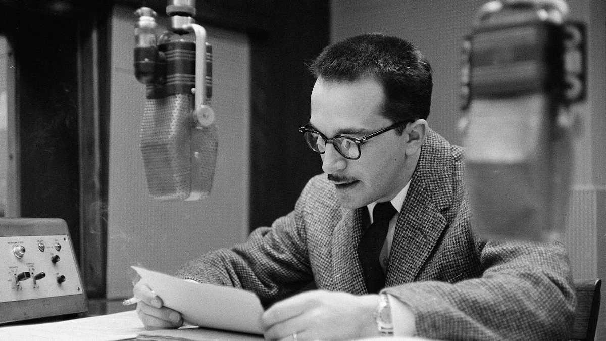 Raymond Charette lisant ses notes dans un studio de radio, derrière le micro.