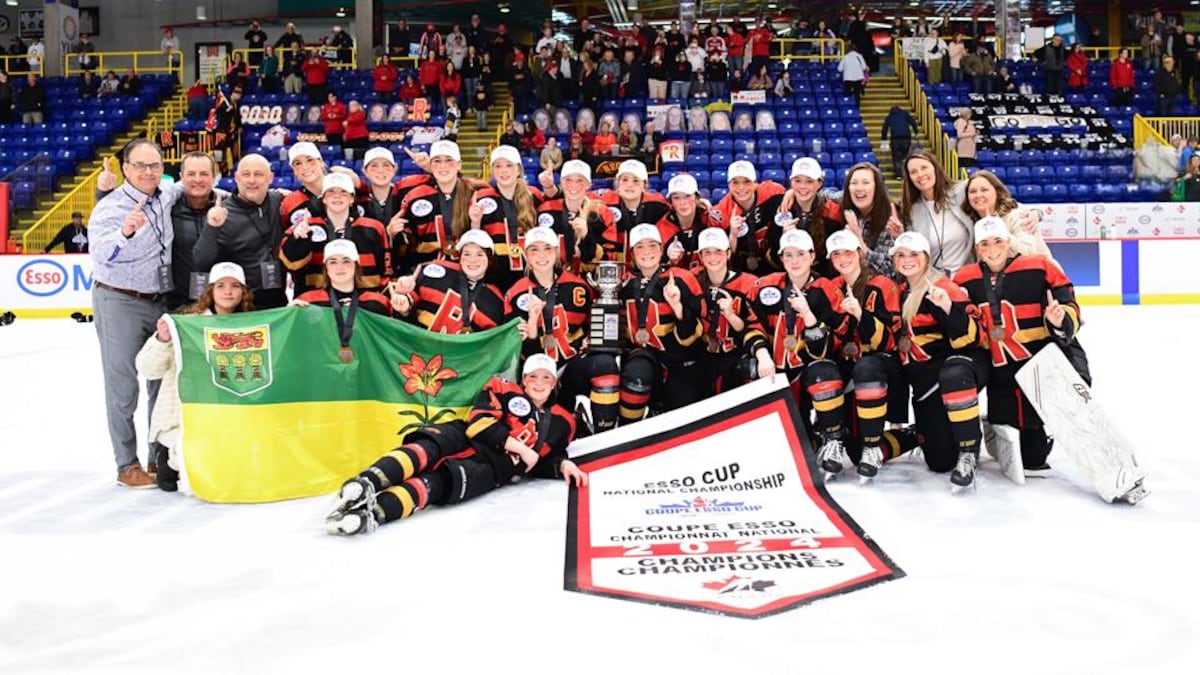 Les joueuses des Rebels de Regina, vêtues de leurs chandails aux couleurs noir, jaune et rouge, posent avec leur bannière de championnes et le drapeau de la Saskatchewan.
