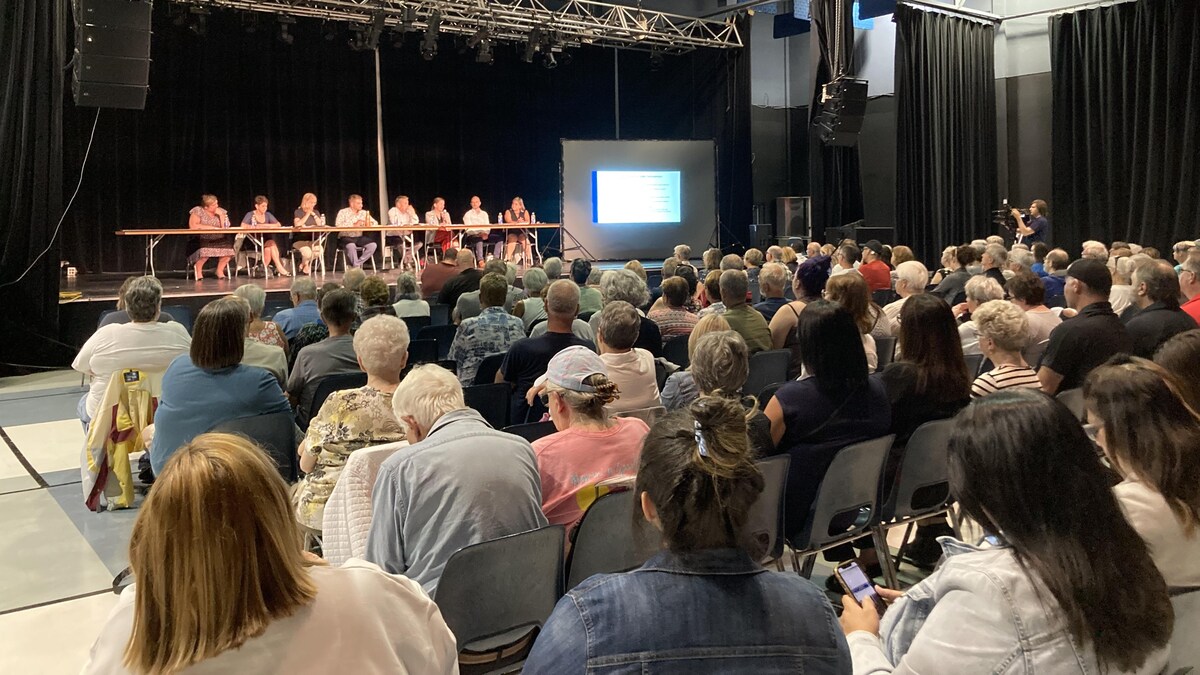 Une centaine de personnes sont assises dans une salle. Devant elles, huit personnes présentent les ruptures de services en santé.