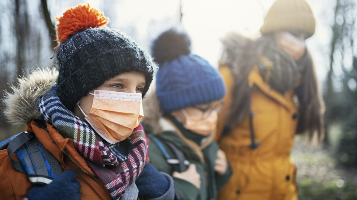 Trois jeunes d'âges différents marchent vers l'école avec des sacs à dos. Ils portent chacun un masque non médical et sont habillés pour le temps froid avec un manteau et une tuque.