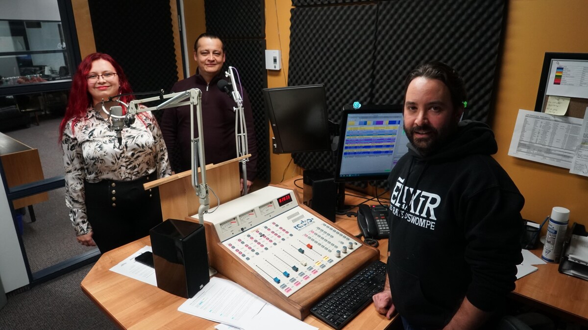 Une femme et deux hommes debout derrière des micros dans un studio d'enregistrement radio.