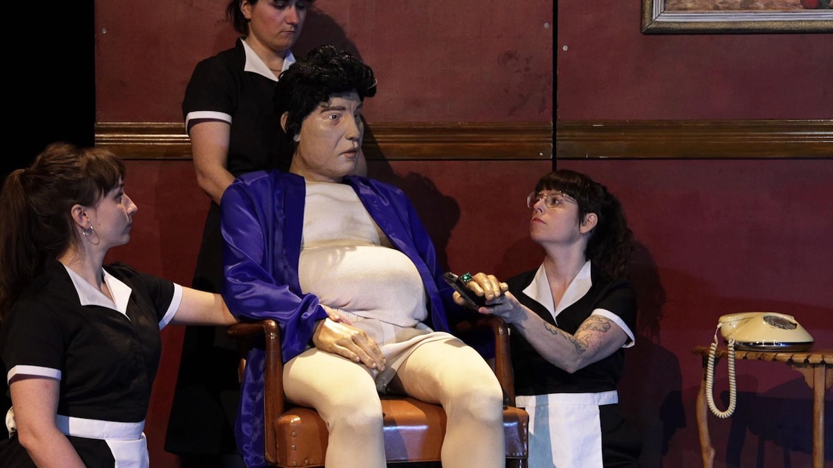 Une scène du spectacle Disgraceland avec trois comédiennes entourant une marionnette géante sur une chaise. 