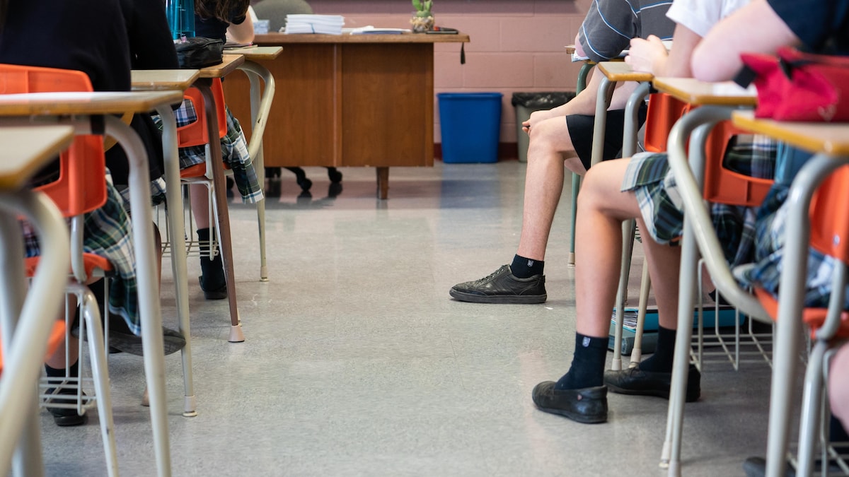 Des élèves du secondaire vus de dos dans une classe à la hauteur des jambes et des pieds, de manière à ne pas pouvoir les identifier.