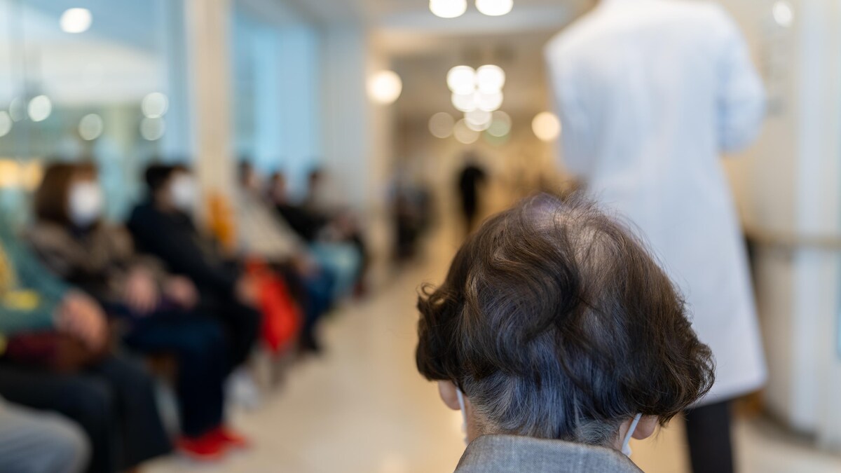 Le derrière de la tête d'une femme âgée dans une salle d'attente. Autour d'elle, on voit les contours flous de nombreuses personnes assises sur des chaises et d'un individu qui passe, vêtu d'un sarrau blanc.