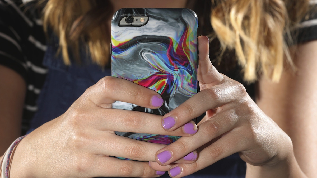 Une jeune femme aux ongles vernis de rose tient devant elle un téléphone cellulaire protégé par une coquille colorée.