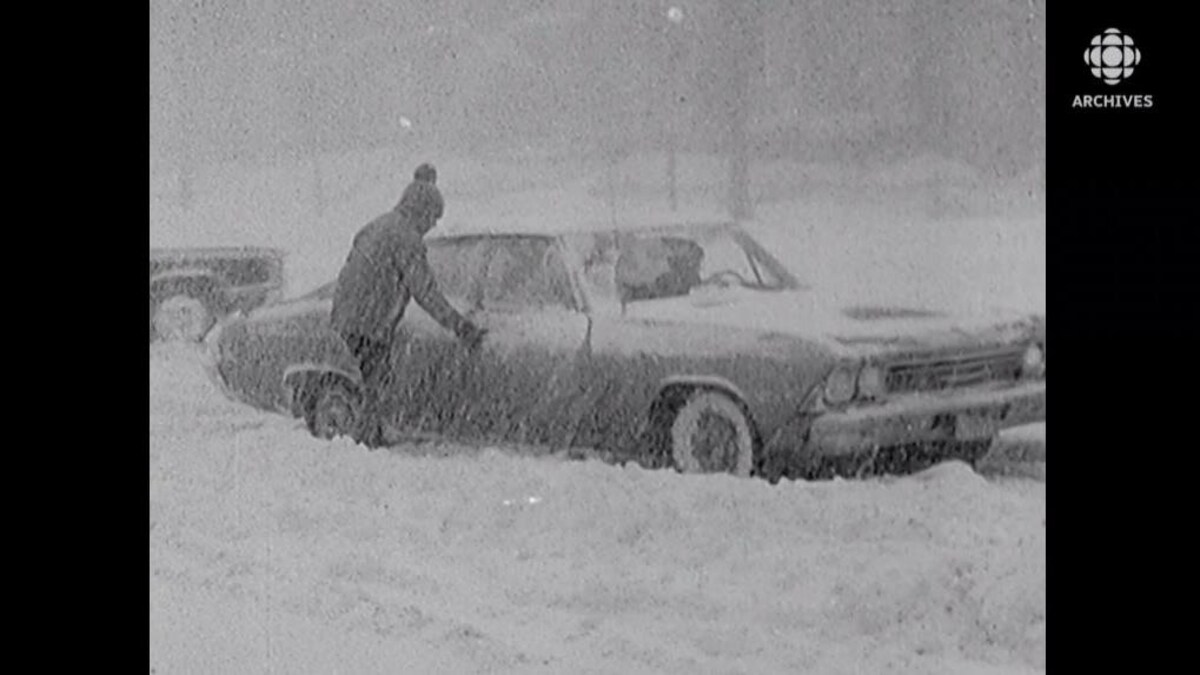 Une personne, sous les flocons, pousse une voiture prise dans un banc de neige